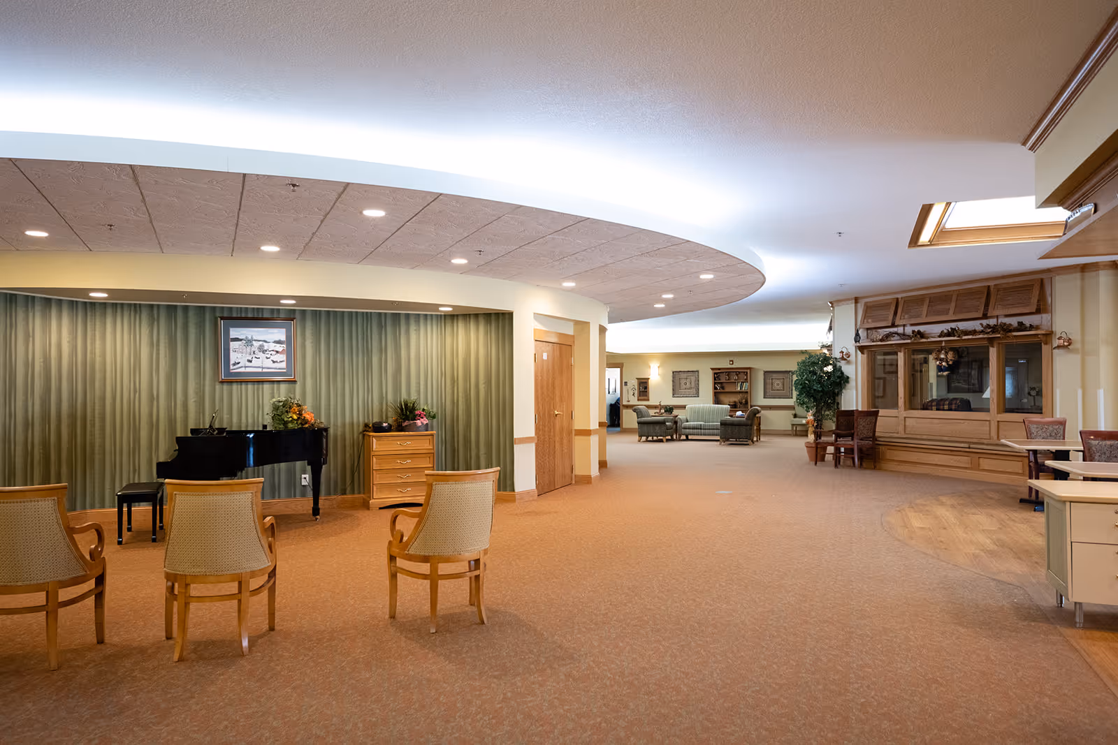 A spacious senior living facility common area with a grand piano, three wooden chairs facing the piano, a wooden chest with flowers on top, and a seating area with sofas and chairs in the background. The ceiling features recessed lighting and a curved design with soft lighting. There is also a wooden display cabinet and a small table with chairs on the right side.