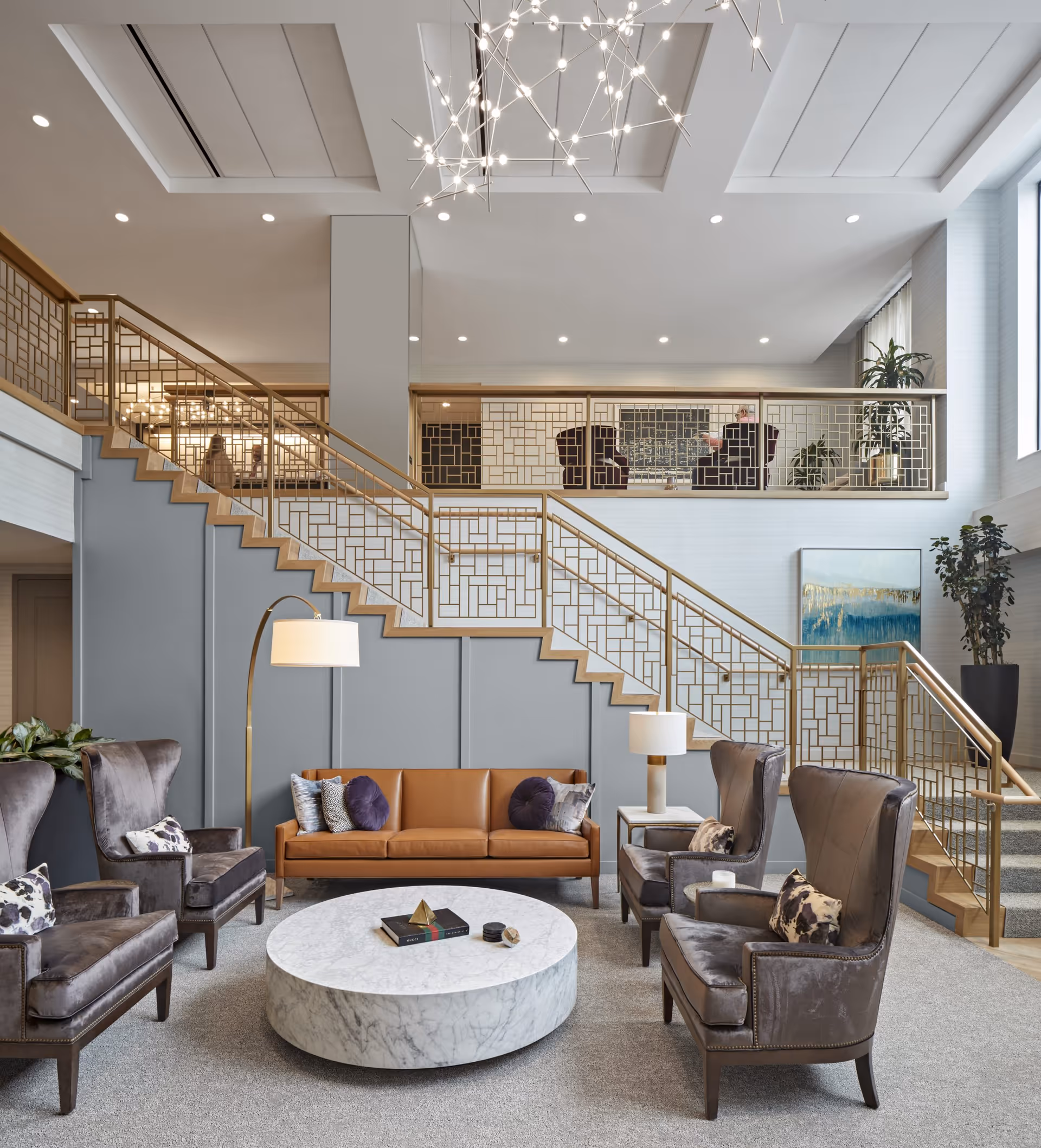A modern and elegant living room area with a brown leather sofa, four gray velvet armchairs with patterned pillows, a round white marble coffee table with decorative items, two floor lamps, and a staircase with gold geometric railings leading to an upper level with additional seating and large windows letting in natural light.