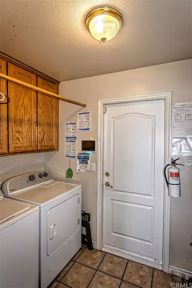 Laundry room with a white washing machine and dryer side by side, wooden cabinets above, a white door, a fire extinguisher mounted on the wall, and a ceiling light fixture.