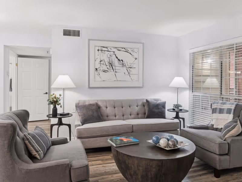 A modern living room with a gray tufted sofa and two matching armchairs arranged around a round dark coffee table. The coffee table has decorative spheres and a book on it. Two side tables with lamps flank the sofa. A large abstract black and white painting hangs on the wall above the sofa. A window with blinds is on the right side, letting in natural light. The floor is wood, and the walls are painted white.