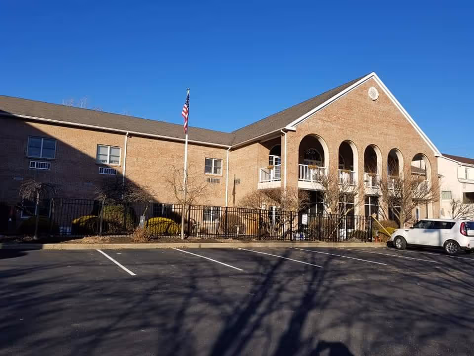 Brick senior living building with arched balconies, an American flag, gated entrance and mostly empty parking lot under a clear blue sky.