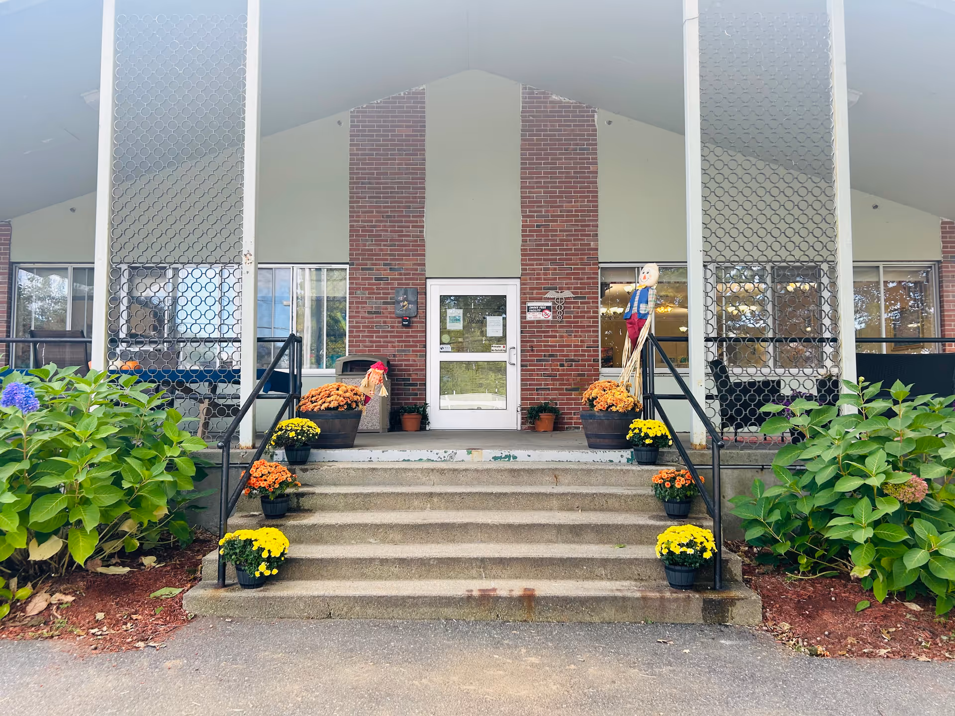 Front entrance of a building with a brick facade and a white door, flanked by large windows. There are concrete steps leading up to the door with black metal railings on both sides. The steps are decorated with multiple pots of yellow and orange flowers. Green bushes are on either side of the steps, and there are two scarecrow decorations near the door.