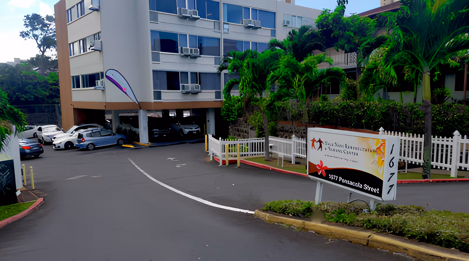 Exterior view of a multi-story building with several parked cars underneath a covered parking area. There are palm trees and greenery around the driveway, and a white fence near a sign displaying the address 1671 and some partially visible text.