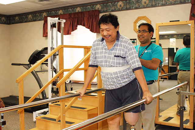 A man with a prosthetic leg is using parallel bars for physical therapy while a therapist stands behind him providing support in a rehabilitation room with exercise equipment and a wooden staircase.