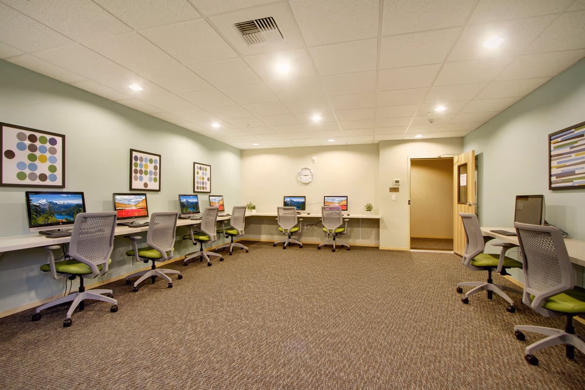 A computer room with multiple workstations along the walls, each equipped with a desktop computer and a green office chair. The room has light green walls, framed abstract artwork, a clock on the far wall, and a carpeted floor. There is a door open on the right side of the room.