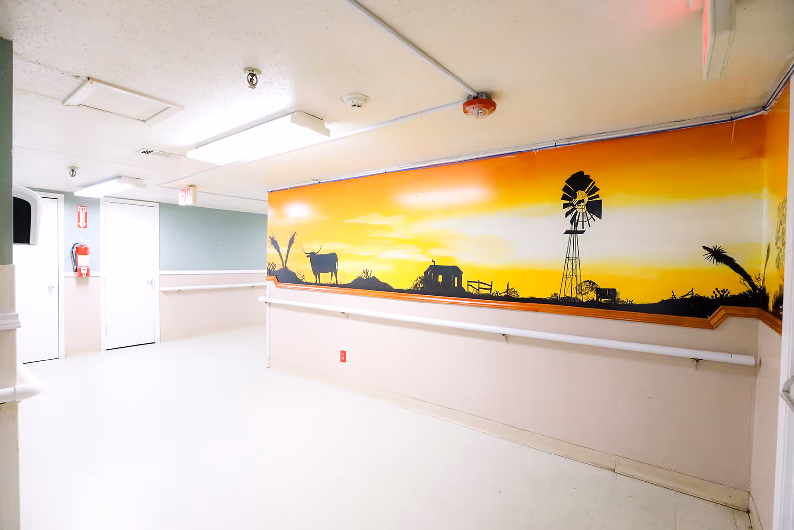 Bright, empty interior corridor with handrails and a large yellow-orange mural of rural silhouettes (windmill, cow, house) on one wall.
