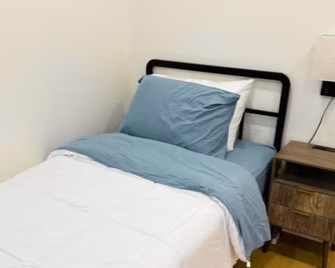 A single bed with blue sheets and a white comforter beside a wooden nightstand and lamp in a simple bedroom.