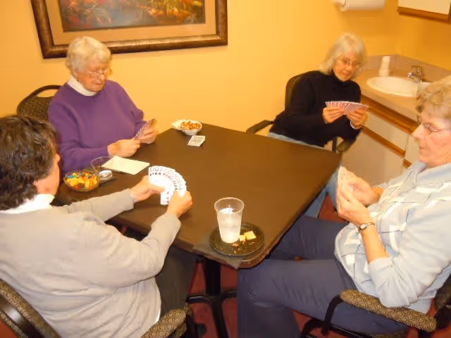 Four elderly women sitting around a square table playing cards in a room with yellow walls. There are snacks and a glass of water on the table, and a framed painting on the wall behind them.
