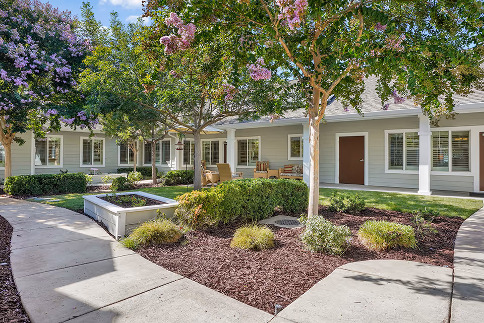Outdoor courtyard area at The Commons on Thornton featuring a curved concrete walkway, landscaped garden beds with mulch, shrubs, and flowering trees. The building exterior is light gray with white trim, multiple windows with white shutters, and brown doors. There is outdoor seating with chairs and cushions under a covered porch area.