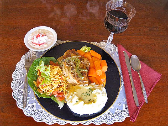 A meal served on a black plate with mashed potatoes topped with gravy and herbs, cooked carrots, a piece of grilled chicken with sauce, and a side salad with lettuce, tomato, and shredded cheese. Next to the plate is a small bowl of dessert with whipped cream and a red topping, a glass of red wine, and silverware placed on a folded maroon napkin on a wooden table with a lace placemat.