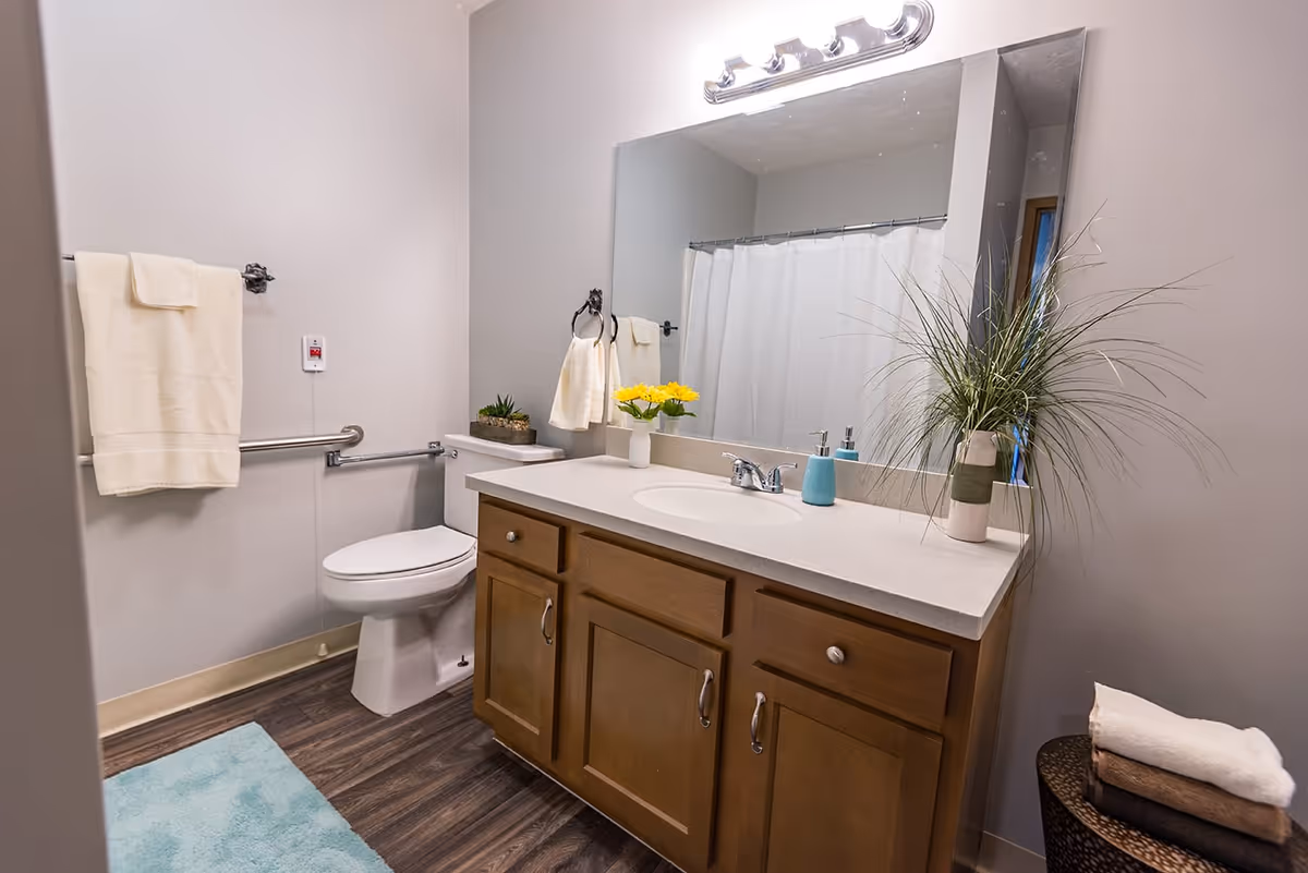 A clean and well-lit bathroom featuring a wooden vanity with a white countertop, a large mirror with lights above, a toilet with a grab bar, a towel rack with a cream-colored towel, a blue bath mat on the floor, and decorative plants and flowers on the countertop and in a basket.