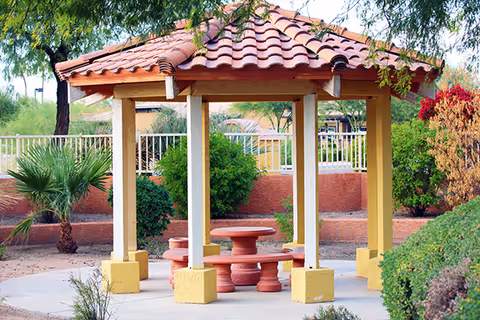 A small outdoor gazebo with a red tiled roof supported by yellow pillars, surrounded by greenery including bushes, small trees, and desert plants. Inside the gazebo is a round red table with matching curved benches. A metal fence and a red brick wall are visible in the background.