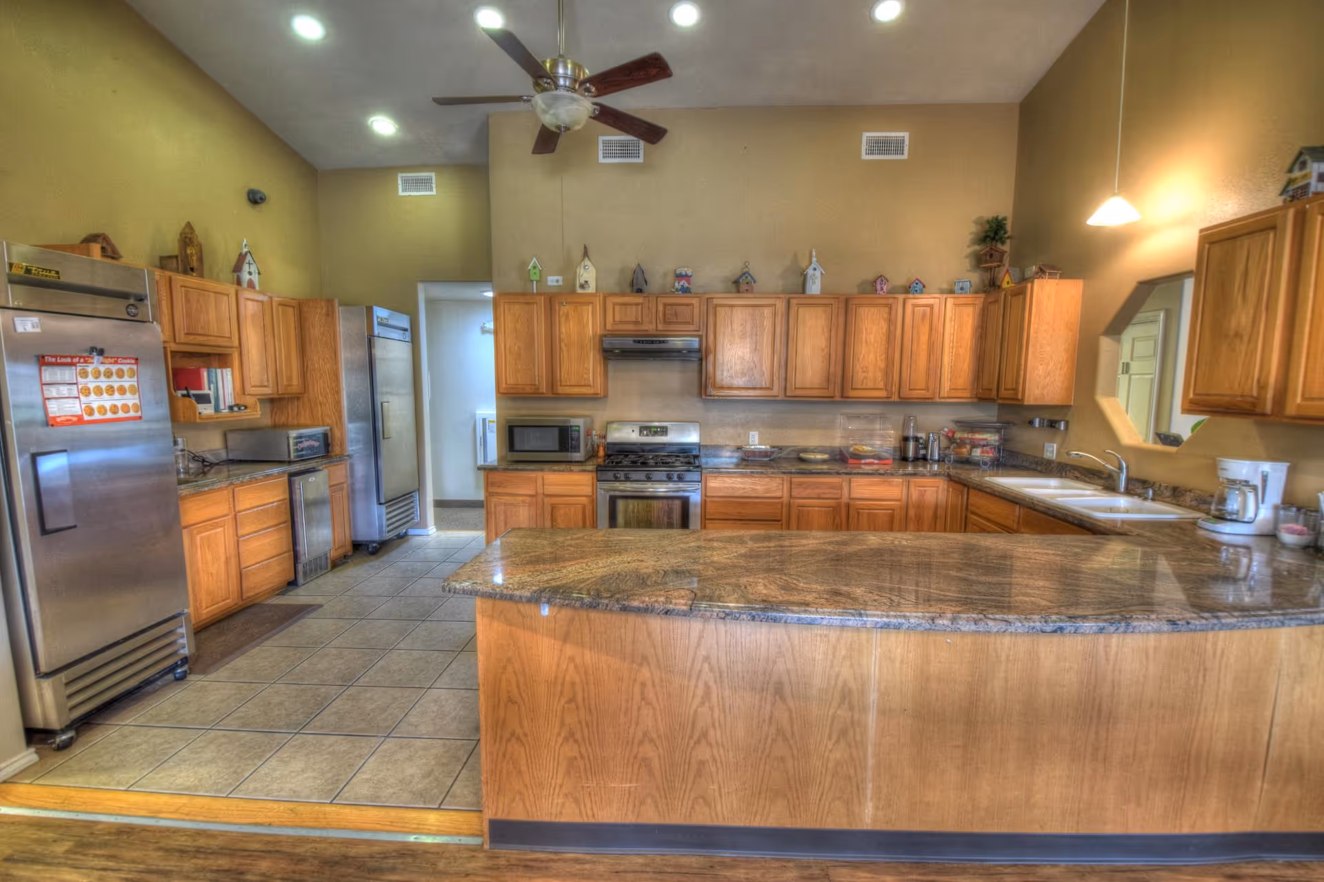 A spacious kitchen with wooden cabinets, granite countertops, and tiled flooring. The kitchen features a large island in the center, a stainless steel refrigerator, a microwave, a stove with an oven, a double sink, and various small kitchen appliances. There is a ceiling fan with lights above and decorative birdhouses on top of the cabinets.