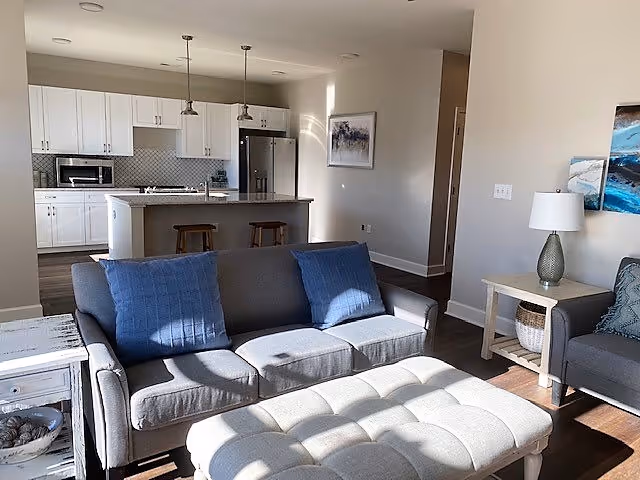 Open-concept living room with a gray sofa, blue pillows and a tufted ottoman facing a kitchen island with pendant lights.