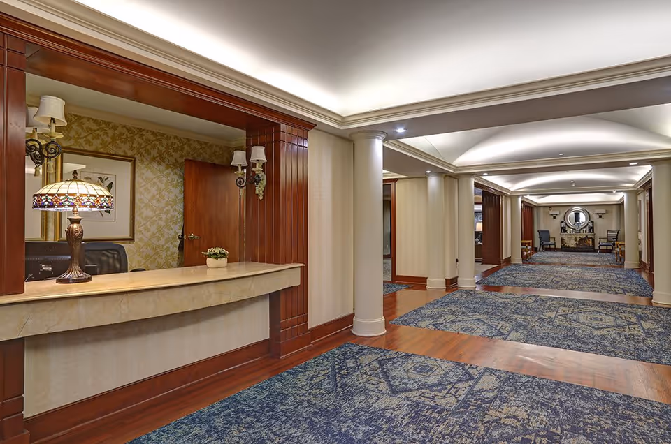 Interior view of a senior living facility hallway with a reception desk on the left featuring a decorative lamp and potted plant. The hallway has blue patterned carpets, wooden flooring accents, cream-colored walls, and white columns. At the far end, there is a seating area with chairs and a round mirror above a fireplace.