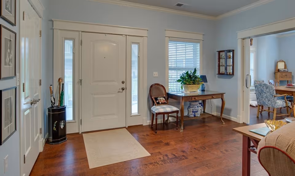Interior view of a senior living facility entrance area with a white front door flanked by narrow windows. To the right, there is a wooden table with a potted plant and a chair with a cushion. The floor is wooden, and the walls are painted light blue. An adjacent room with dining chairs and a table is visible through an open doorway.
