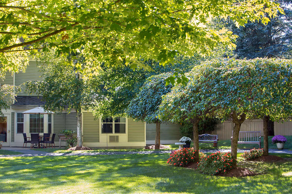 A peaceful outdoor garden area at Benchmark Senior Living at Putnam Farm featuring well-maintained green grass, several small trees with dense foliage, colorful flower beds, benches, and a patio with a table and chairs under an umbrella near the building.