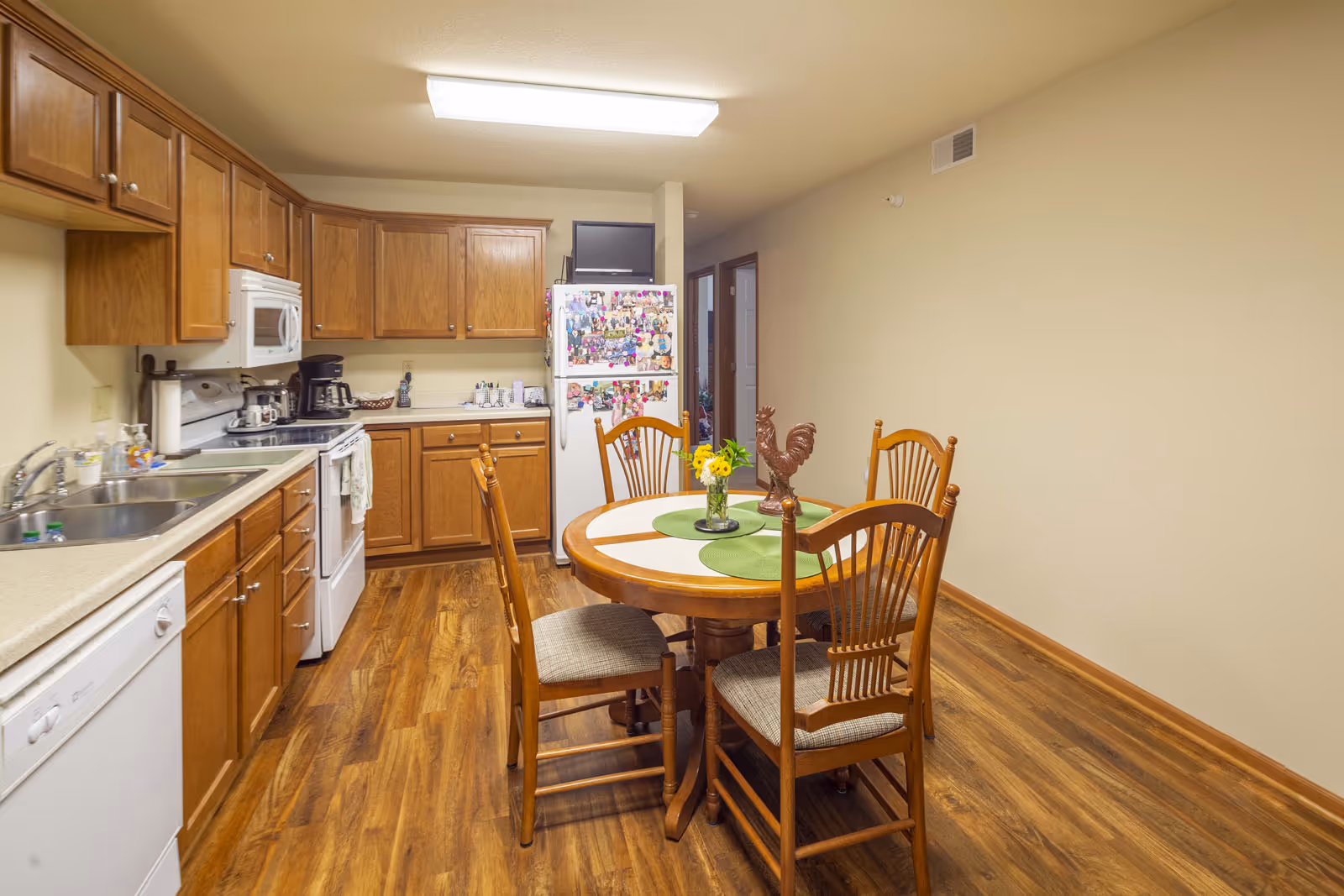 A kitchen and dining area with wooden cabinets, a white stove, microwave, dishwasher, and refrigerator covered with photos and magnets. A round wooden dining table with four chairs is in the center, decorated with a small vase of yellow flowers and a rooster figurine. The floor has wood-style flooring and the walls are painted beige.