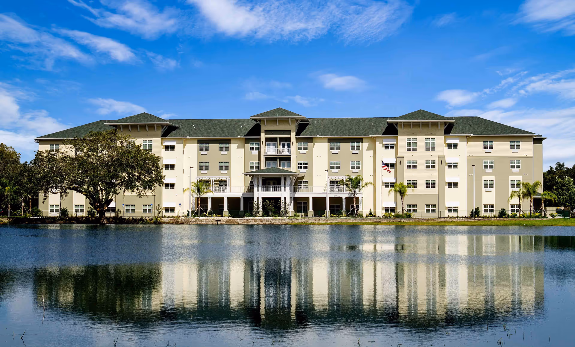 A large, multi-story senior living facility building with a green roof and beige exterior, reflected in a calm body of water in front. The building has many windows and a central entrance with a covered porch. There are trees and palm plants around the building under a partly cloudy blue sky.