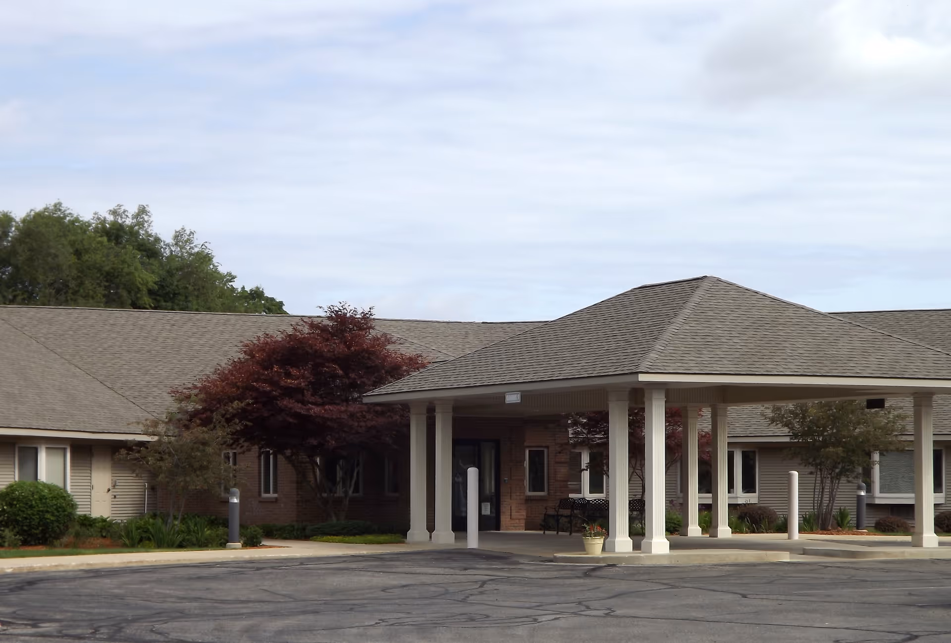 Exterior view of Railside Assisted Living Center showing the entrance with a covered drop-off area supported by white columns, surrounded by trees and shrubs under a cloudy sky.