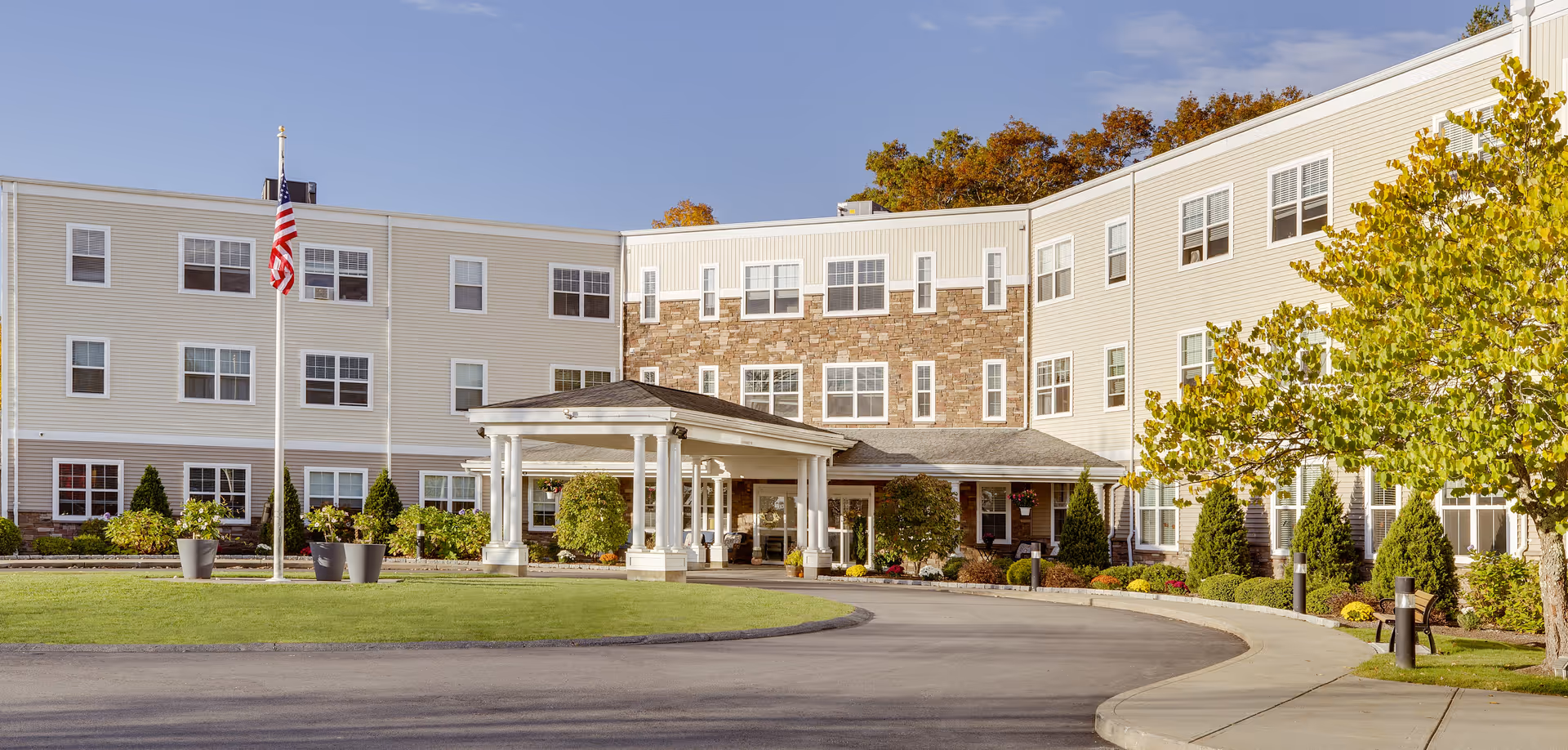 Exterior view of a senior living facility named The Residence at Great Woods, showing a three-story building with beige siding and stone accents. There is a covered entrance with white columns, a curved driveway, well-maintained landscaping, an American flag on a flagpole, and trees with autumn foliage under a clear blue sky.
