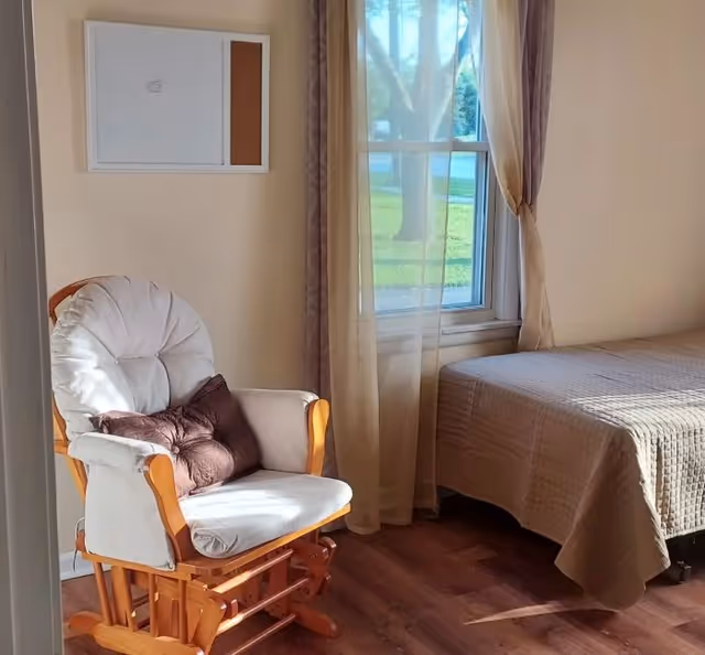A cozy room with a cushioned wooden rocking chair with two brown pillows, a bed with a beige quilt, a window with sheer curtains letting in natural light, and a bulletin board on the wall.