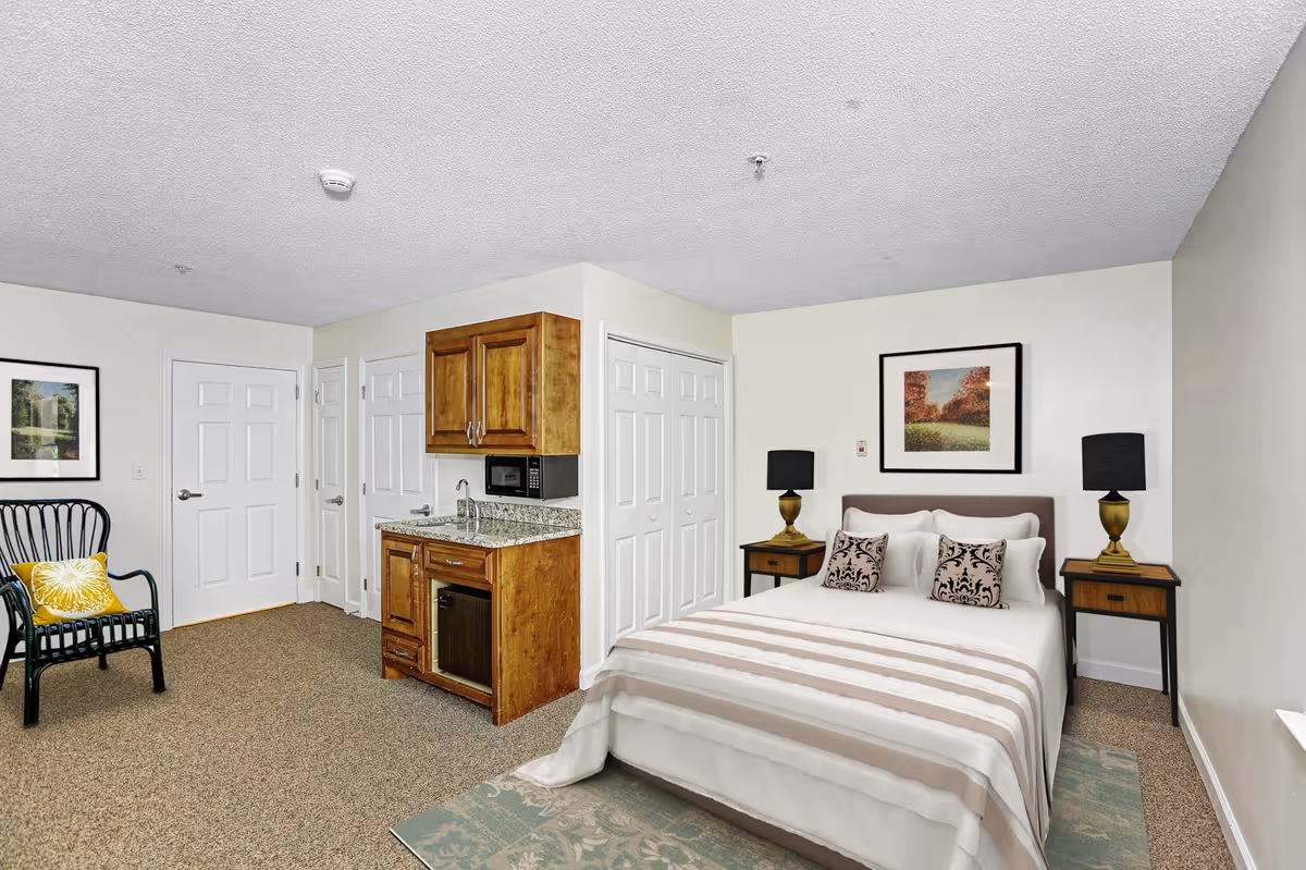 A cozy bedroom in a senior living facility featuring a neatly made bed with striped bedding and decorative pillows, two bedside tables with matching lamps, a small kitchenette area with wooden cabinets, a microwave, and a mini fridge. There is a black chair with a yellow cushion, framed artwork on the walls, and a carpeted floor.