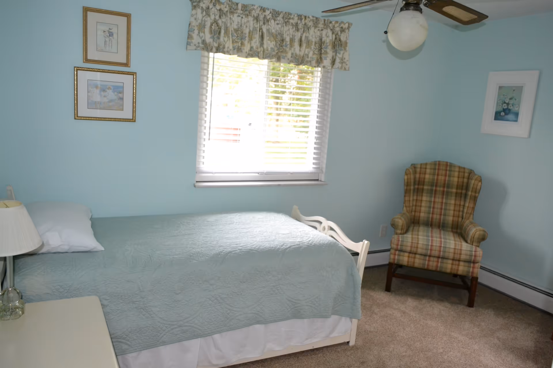 Small bedroom with a single bed covered in a light blue quilt, a window with blinds, and a plaid armchair.