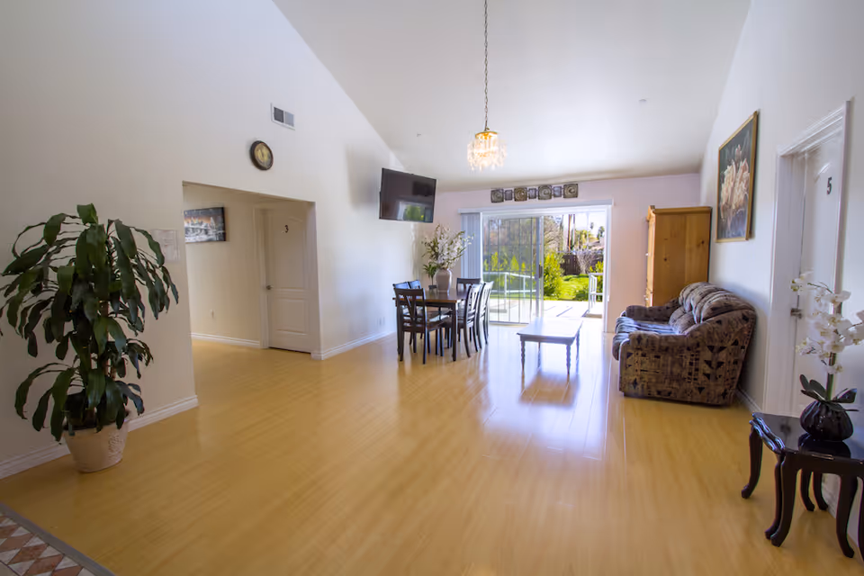 A bright and spacious living area in an assisted living facility with light wooden flooring, a dining table with chairs, a wall-mounted TV, a patterned sofa, a coffee table, and large sliding glass doors leading to a green outdoor garden. There are doors labeled with numbers 3 and 5, a large potted plant, a small side table with a flower vase, and wall decorations including a clock and framed artwork.