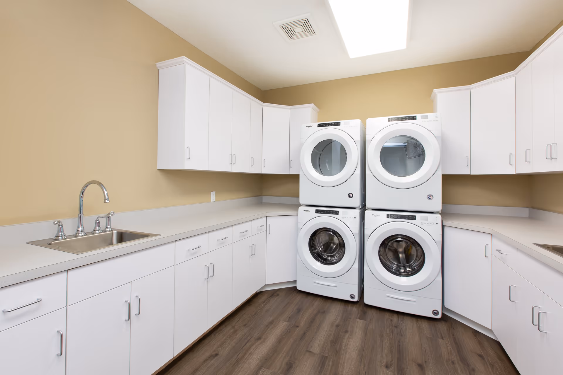 A clean laundry room with two stacked sets of white Whirlpool front-loading washers and dryers, white cabinets above and below a long countertop, a stainless steel sink with a chrome faucet, beige walls, and wood-look flooring.