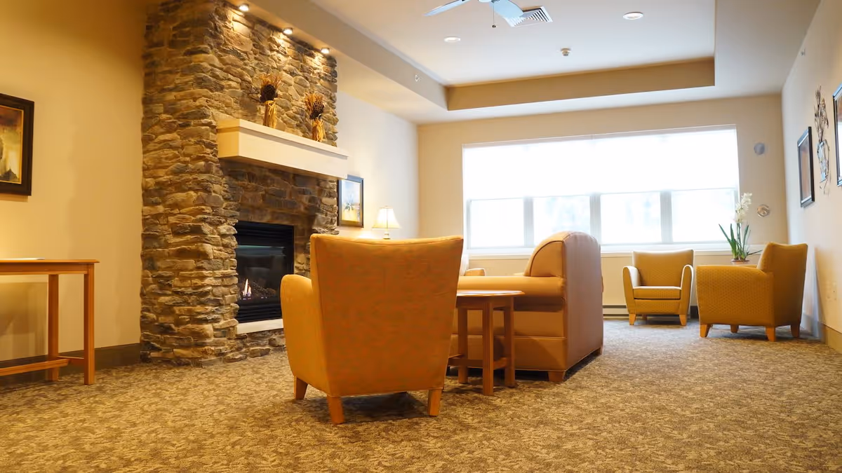 A cozy living room area with a stone fireplace on the left wall, surrounded by warm-toned armchairs and a sofa. There is a large window letting in natural light, a small wooden table, and decorative items on the walls and mantel.