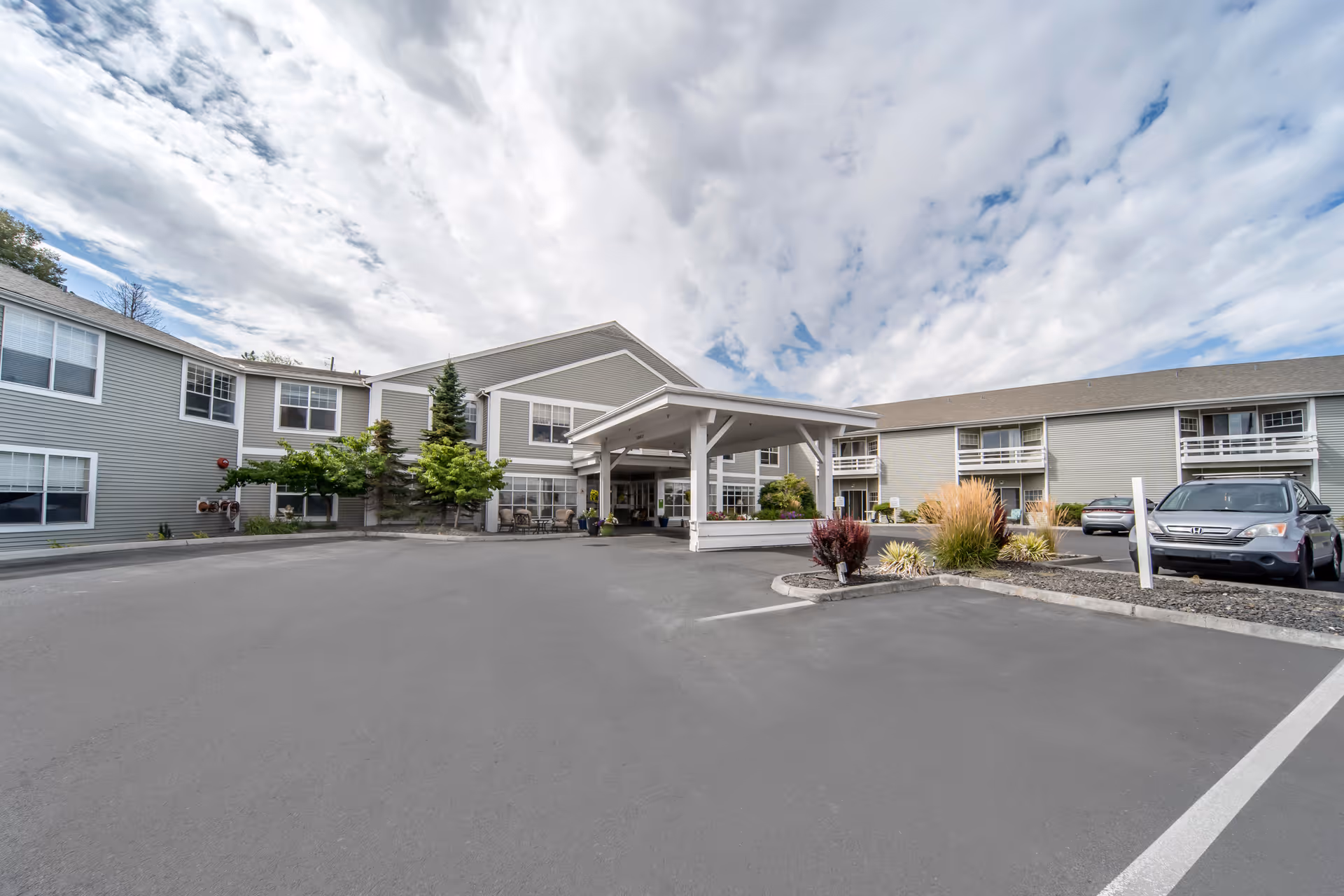 Exterior view of Brookdale Hearthstone Moses Lake senior living facility showing a two-story building with gray siding, multiple windows, a covered entrance with white pillars, a parking lot with several cars, and landscaped areas with shrubs and small trees under a partly cloudy sky.
