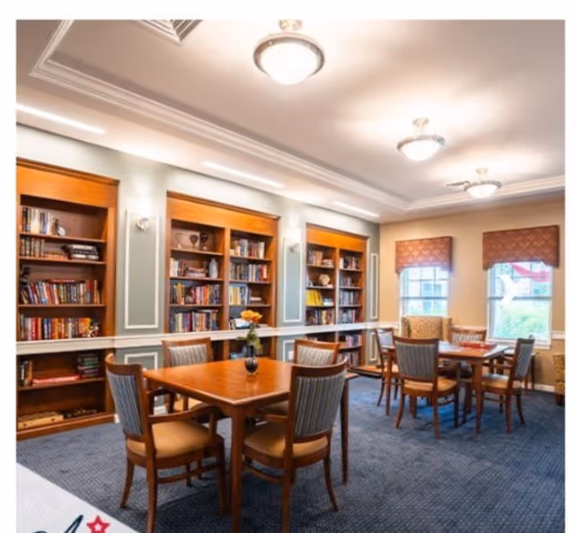 Well-lit common room with wooden tables and chairs and built-in bookshelves along one wall.