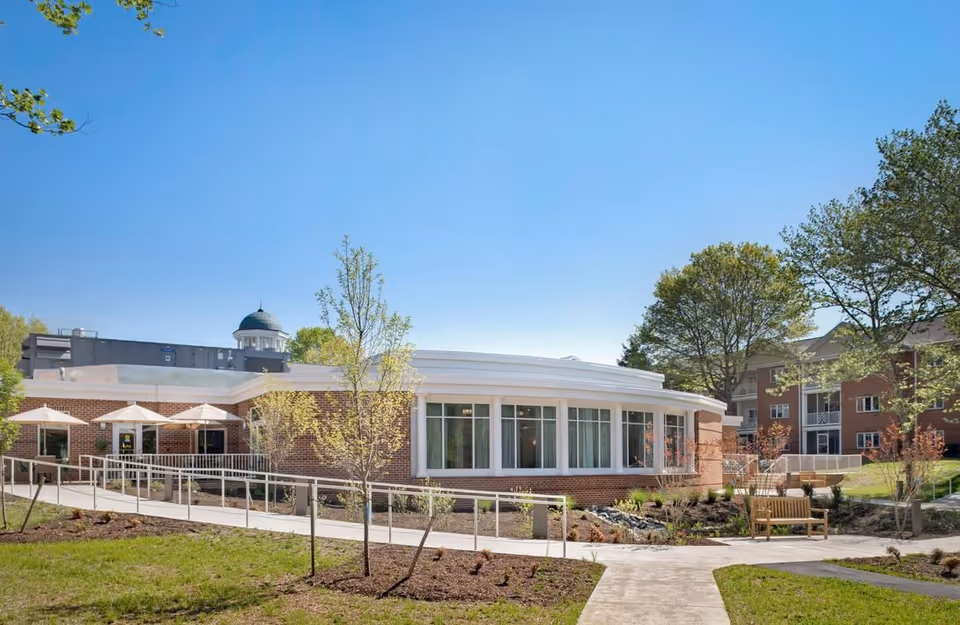 Exterior view of a senior living facility named The Colonnades, featuring a curved brick building with large windows, a ramp with handrails, outdoor umbrellas, benches, and landscaped greenery under a clear blue sky.