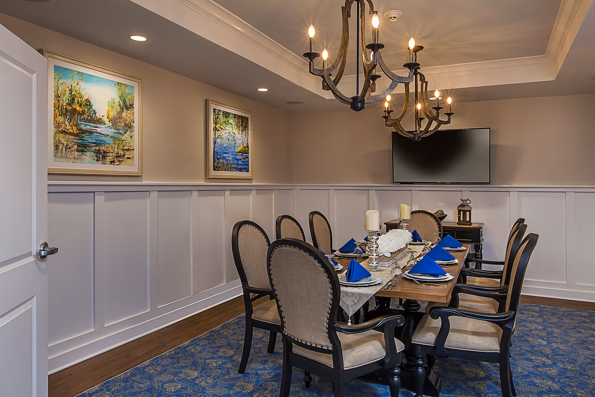 A formal dining room with a wooden table set for eight people. The table is decorated with blue napkins, silver candle holders, and a floral centerpiece. The room features beige walls with white wainscoting, two landscape paintings, a flat-screen TV mounted on the wall, and a chandelier with multiple candle-style lights. The floor has a blue patterned carpet.