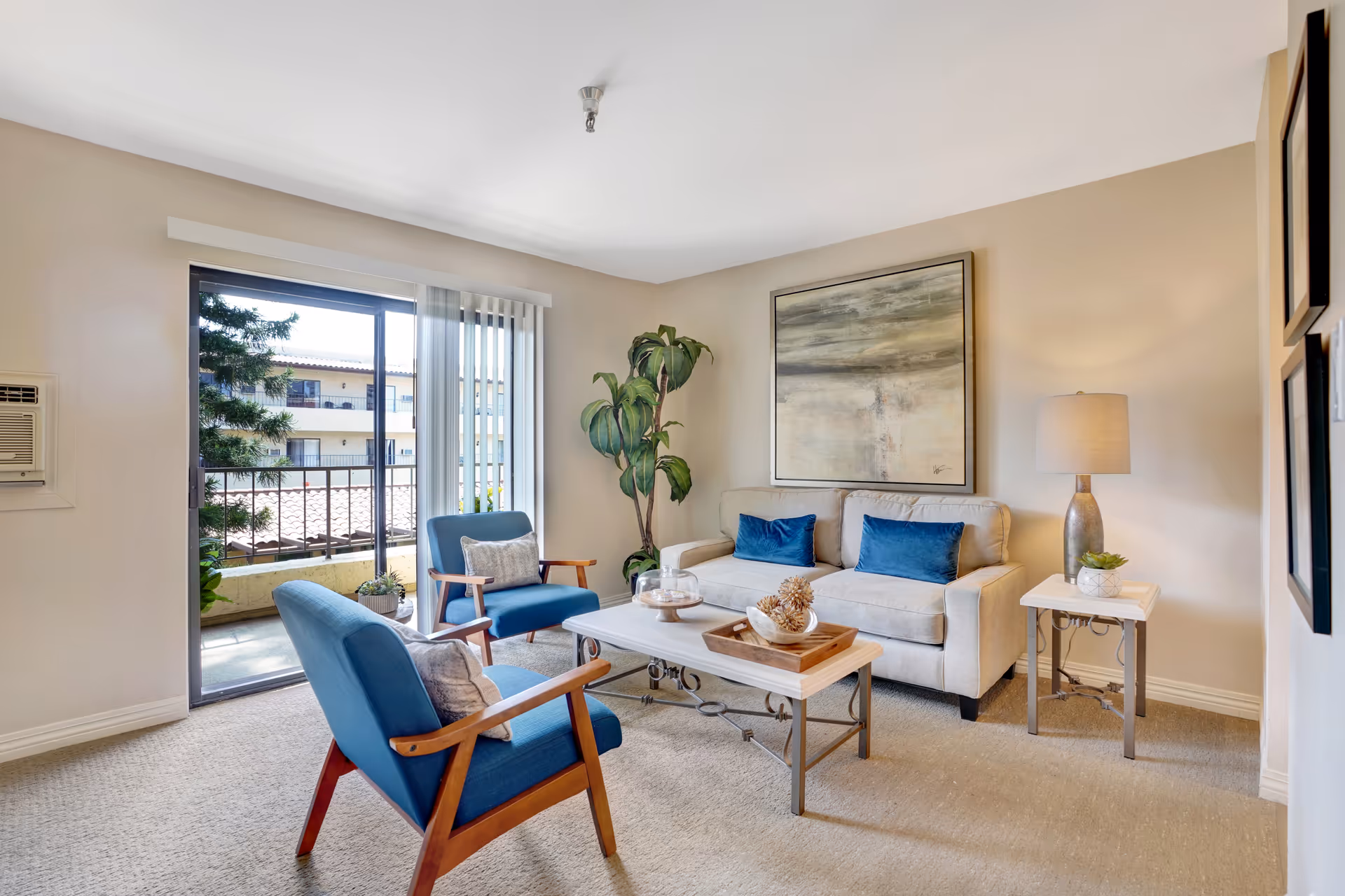 A bright living room with a beige sofa adorned with two blue cushions, two blue armchairs with wooden arms, a coffee table with decorative items, a side table with a lamp and a small plant, a large abstract painting on the wall, and a sliding glass door leading to a balcony with a view of another building and some greenery.