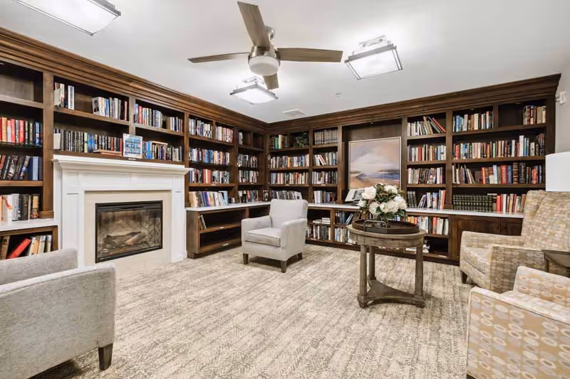 A cozy library room with built-in dark wood bookshelves filled with books, a white fireplace, a round wooden table with a flower arrangement, and several upholstered armchairs arranged around the table. The room has a beige carpet and ceiling lights with a ceiling fan.