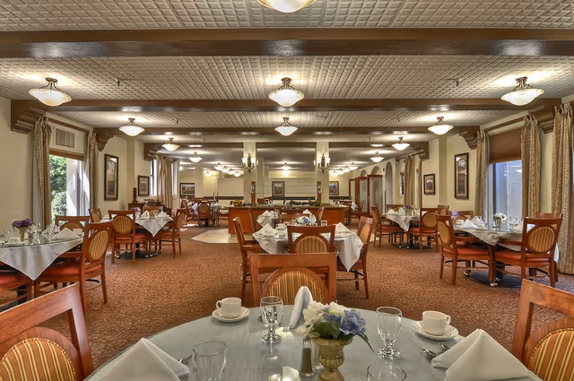 A spacious dining room with multiple round tables covered with white tablecloths, set with cups, glasses, napkins, and small flower arrangements. The room has warm lighting from ceiling fixtures, large windows with curtains, and framed artwork on the walls.