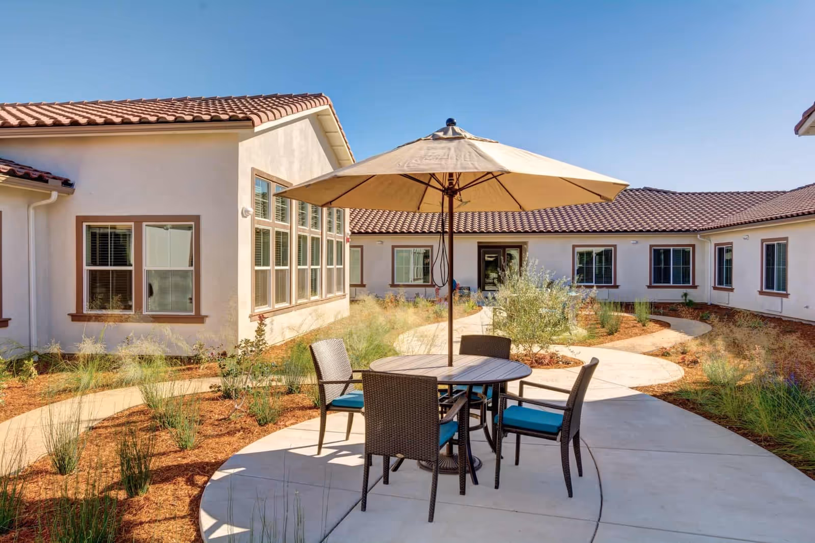 Outdoor courtyard area at Rose Arbor Village featuring a round table with four wicker chairs with blue cushions and a large beige umbrella. The courtyard is surrounded by single-story buildings with white walls and red tile roofs, with a curved concrete pathway and landscaped plants and shrubs under a clear blue sky.
