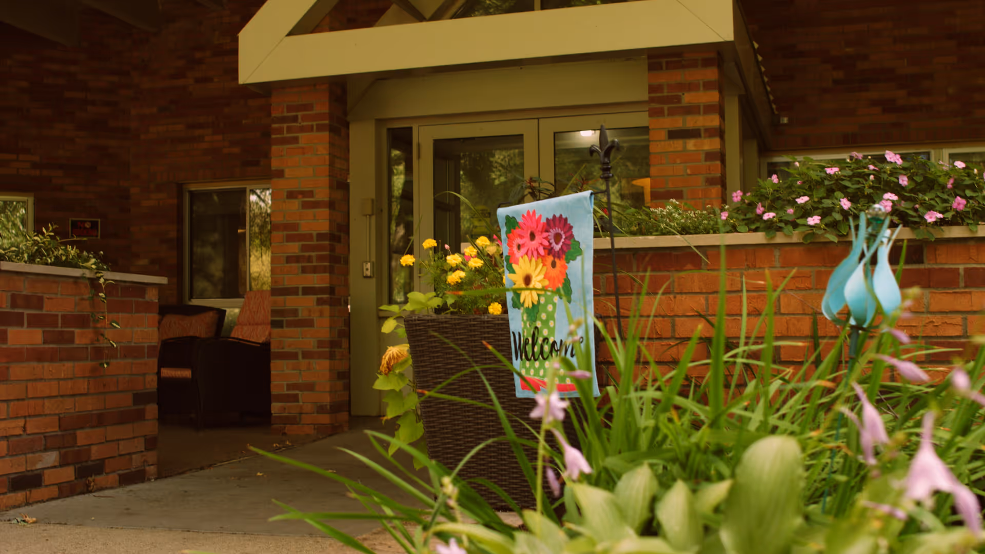 Entrance to a brick building with a covered porch area. There are potted plants and flowers, including a colorful welcome flag with flowers on it. Two chairs are visible inside near the entrance.