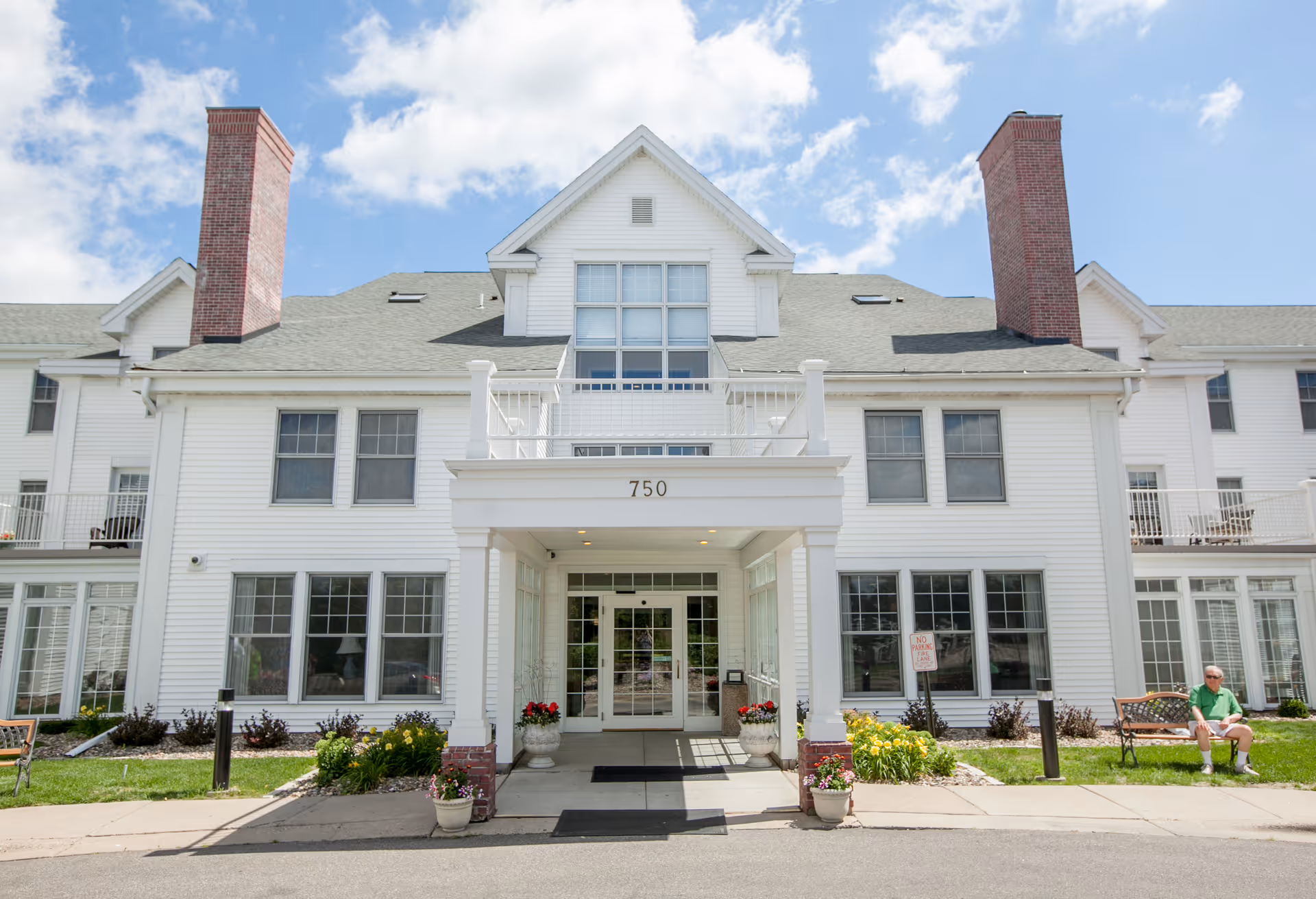 Front entrance of a white senior living building labeled '750' with flower pots, benches and a person sitting on the right.