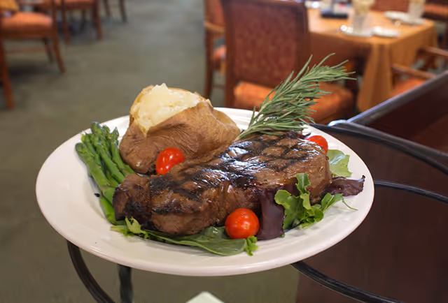 A plate of food featuring a grilled steak garnished with a sprig of rosemary, cherry tomatoes, leafy greens, steamed asparagus, and a baked potato with a split top, served in a dining area with tables and chairs in the background.