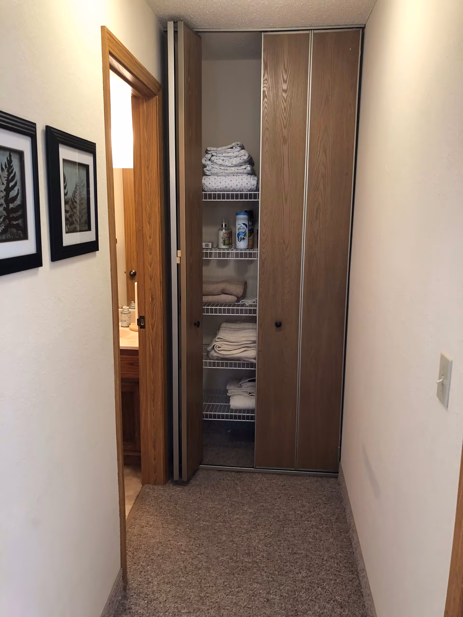 A narrow hallway with beige carpet leading to a small closet with bi-fold wooden doors. The closet contains neatly folded towels and some toiletry items on wire shelves. To the left of the closet is an open door revealing a bathroom sink and mirror. Two framed pictures hang on the left wall of the hallway.