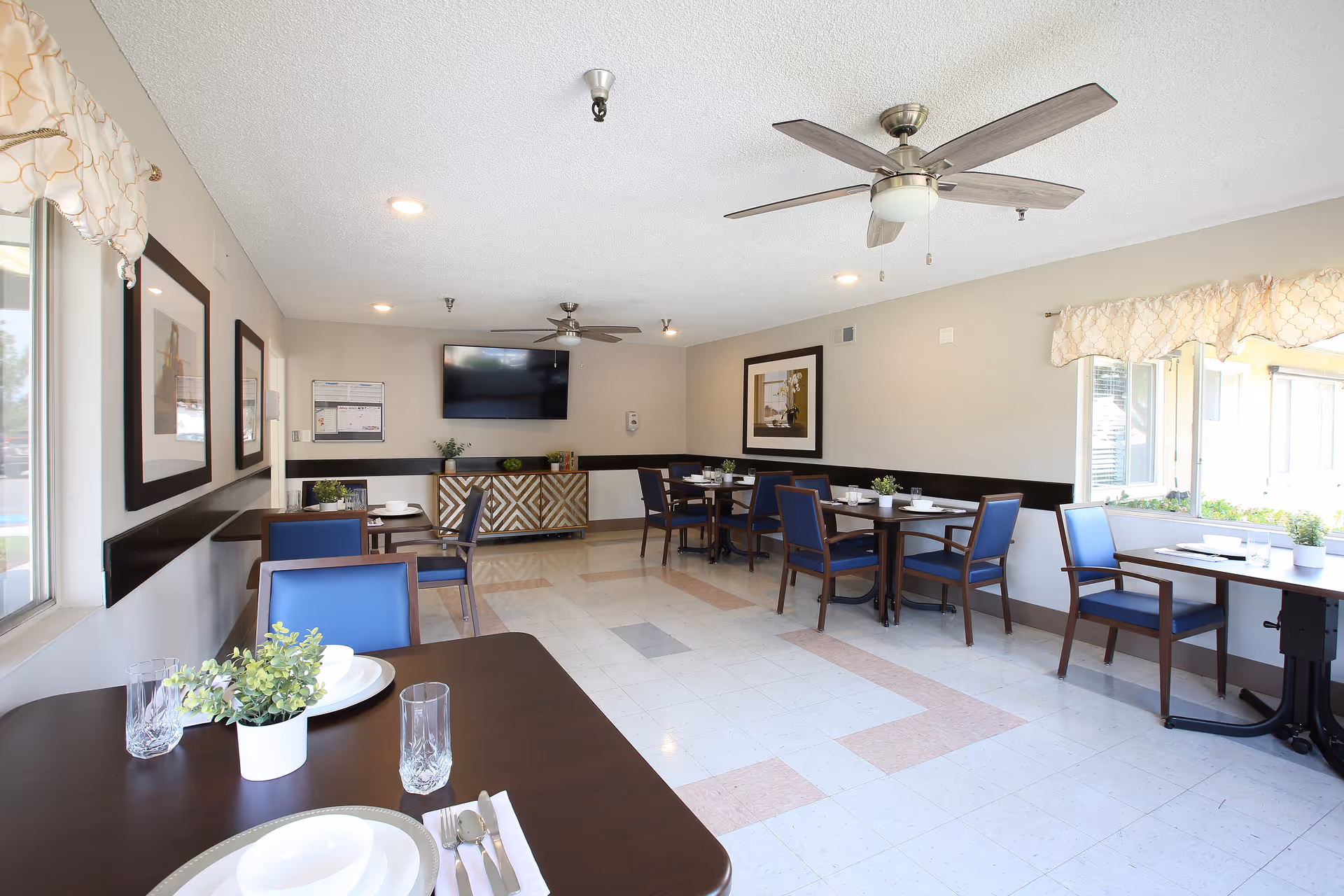 Bright communal dining room with multiple tables and blue chairs, ceiling fans, a wall-mounted TV, and large windows.
