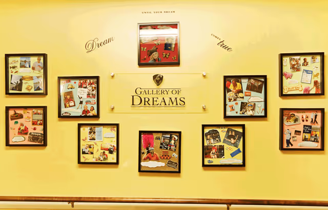 A yellow wall with a display titled 'Gallery of Dreams' featuring nine framed collage boards with various photos and clippings. Above the display, the words 'Until your dream comes true' are written in a curved arrangement.