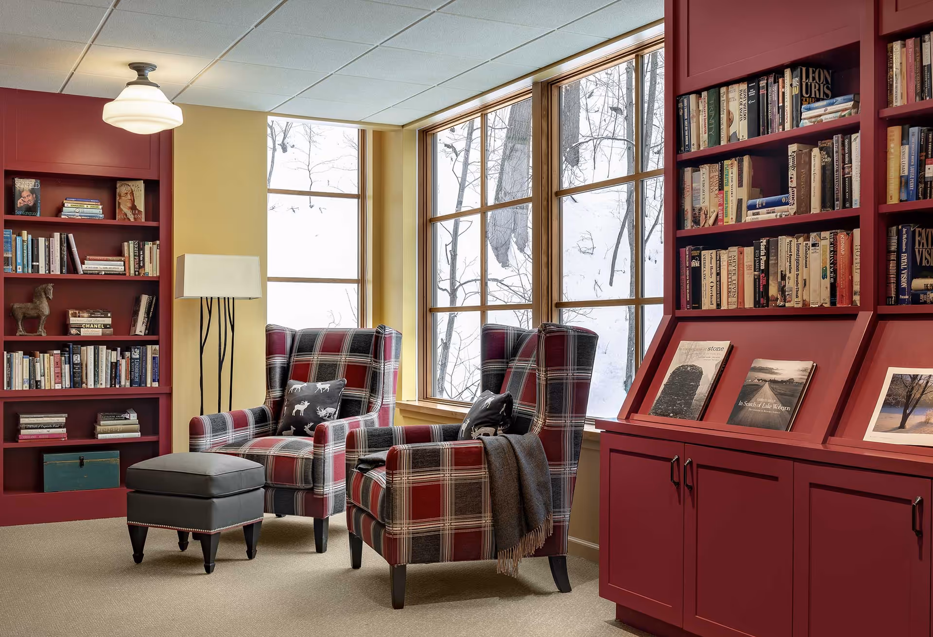 A cozy reading nook with two plaid armchairs and a small ottoman in front of large windows showing a snowy outdoor scene. The room features built-in red bookshelves filled with books and framed pictures, a floor lamp, and beige carpeted flooring.