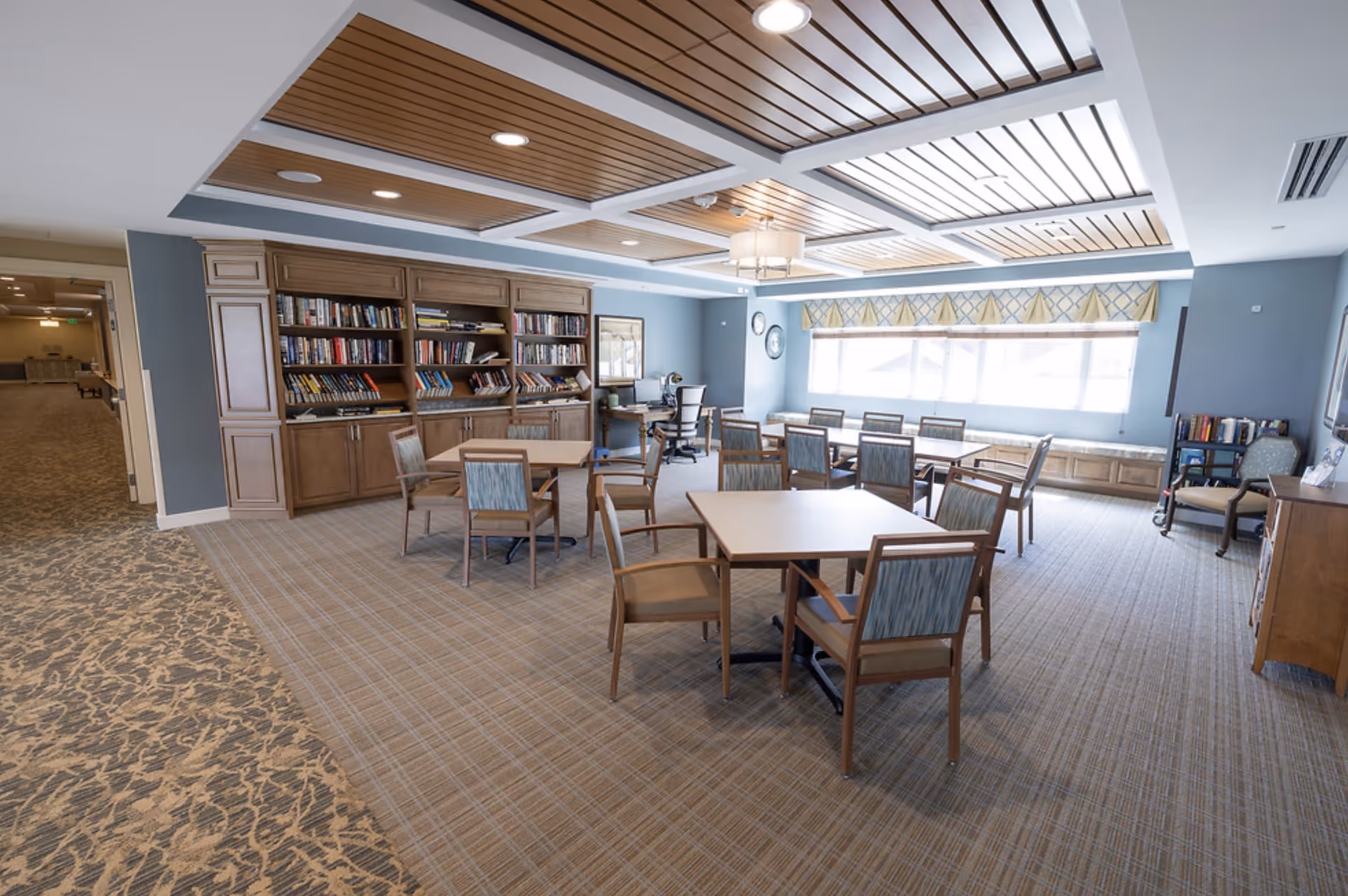 A bright and spacious common area with multiple tables and chairs arranged for seating. The room features a wooden ceiling with recessed lighting, a large bookshelf filled with books, a desk with a computer, and large windows with patterned valances allowing natural light to fill the space. The carpet has a patterned design, and the walls are painted light blue.