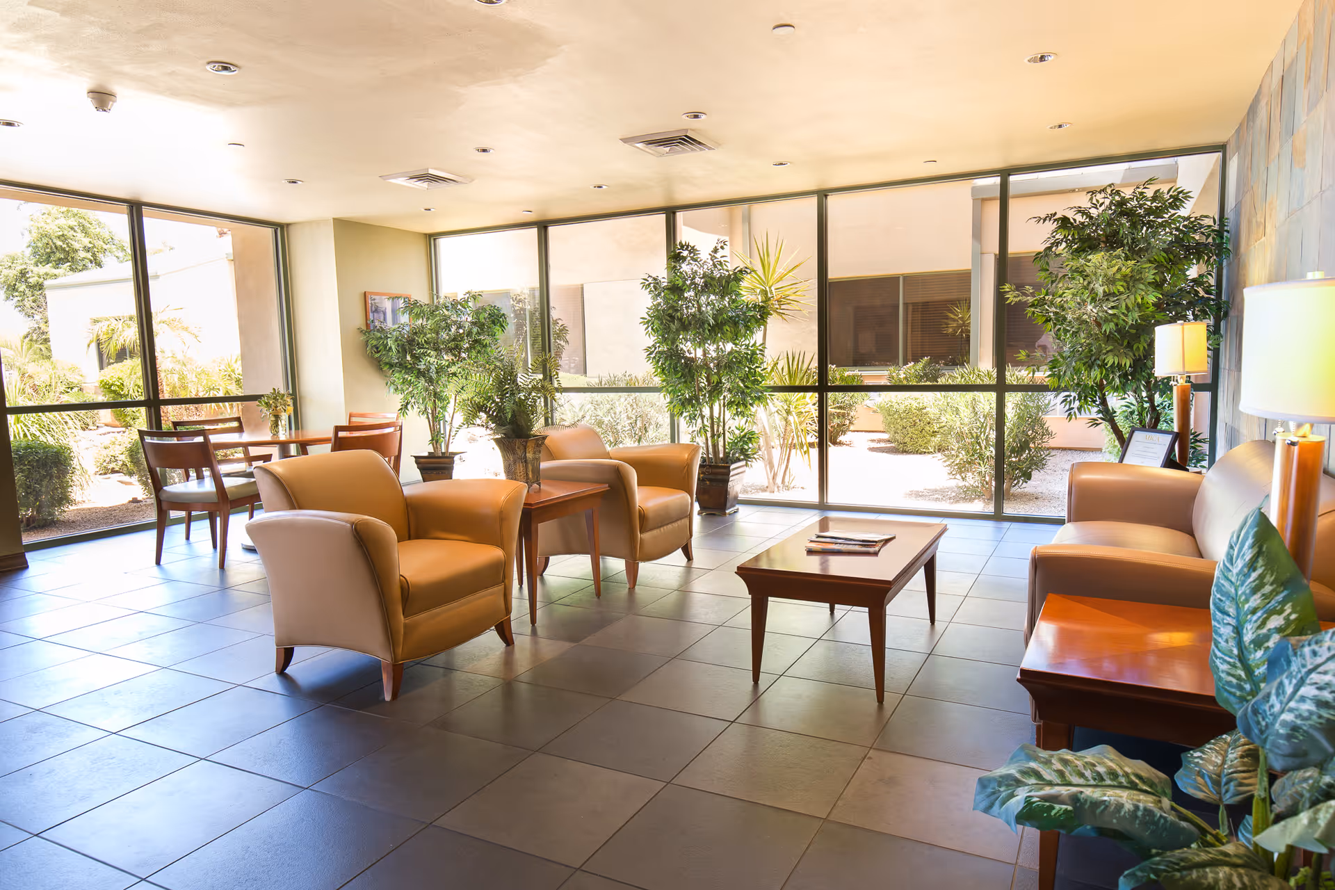 A bright and spacious sitting area with large floor-to-ceiling windows letting in natural light. The room features beige armchairs, a wooden coffee table with magazines, side tables with lamps, and several potted plants. Outside the windows, greenery and bushes are visible.