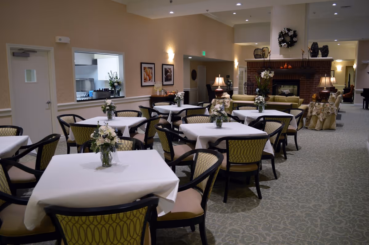 A dining room in a senior living facility with several tables covered in white tablecloths, each adorned with a vase of white flowers. The room features patterned chairs, a carpeted floor, soft lighting, and a brick fireplace with decorative items on the mantel. There is a serving window to a kitchen area on the left wall and framed pictures hanging above a sideboard.