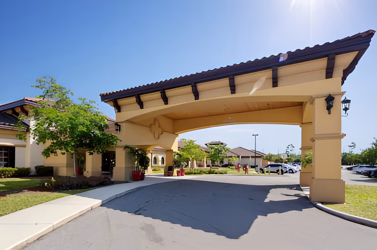 Entrance area of Discovery Commons Cypress Point facility featuring a large covered driveway with a tiled roof, beige columns, and surrounding greenery under a clear blue sky.